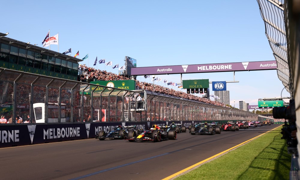 Formula one cars on a race track with promotional signs and a grand stand with a large crowd of people.