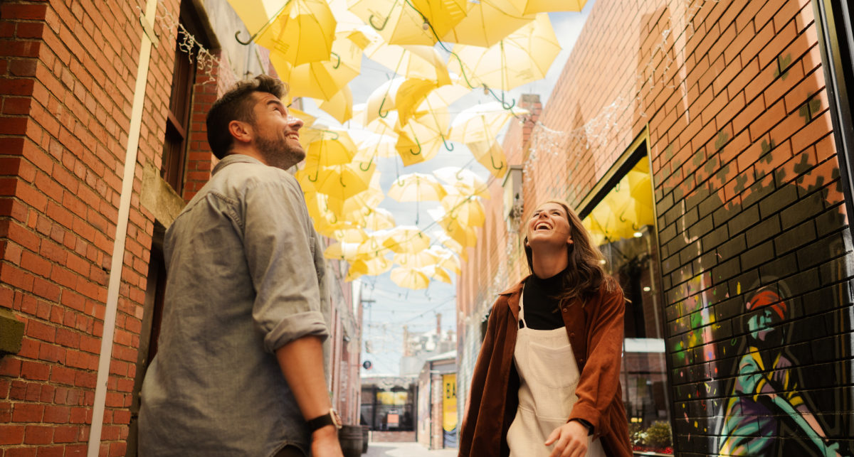 Smiling man and woman in an alleyway looking up at hanging yellow umbrellas.