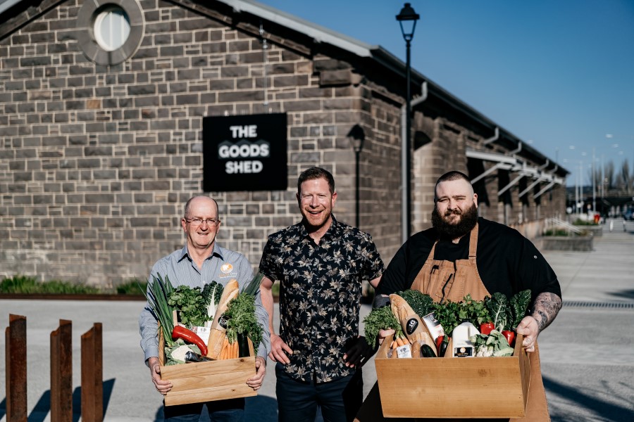 Three people standing out front of The Goods Shed with two holding a box of produce.