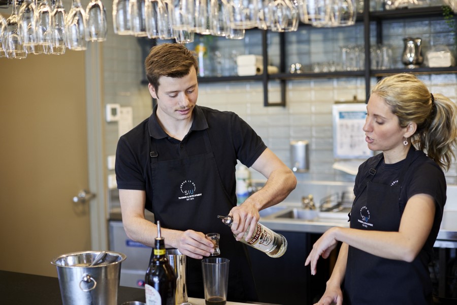 Person measuring an alcoholic drink with colleague guiding.