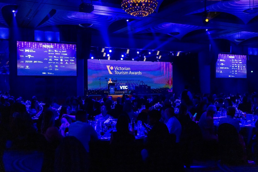 Background screen with Victorian Tourism Awards on projector and person on stage behind lectern,  Foreground people sitting at round tables.  Dimly lit blue with stage lights on presenter. 