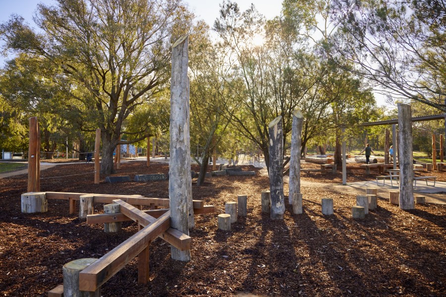 Wooden walking plans in a tambark playground with tall wood stumps among green trees.