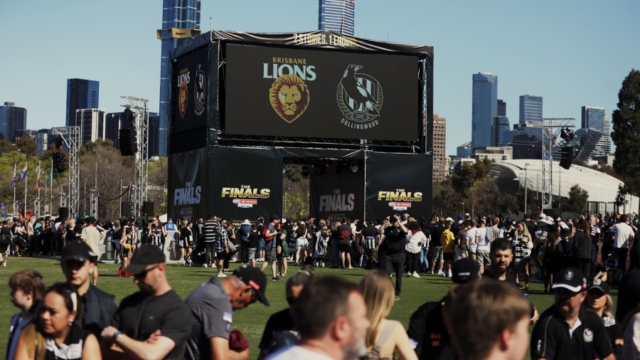 Large crowd standing in front of giant tv screen in park with Brisbane Lions and Collingwood logo.